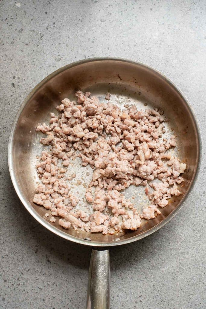 A frying pan on a gray countertop containing partially cooked ground meat, slightly browned in spots. The meat is scattered unevenly across the pan's surface.