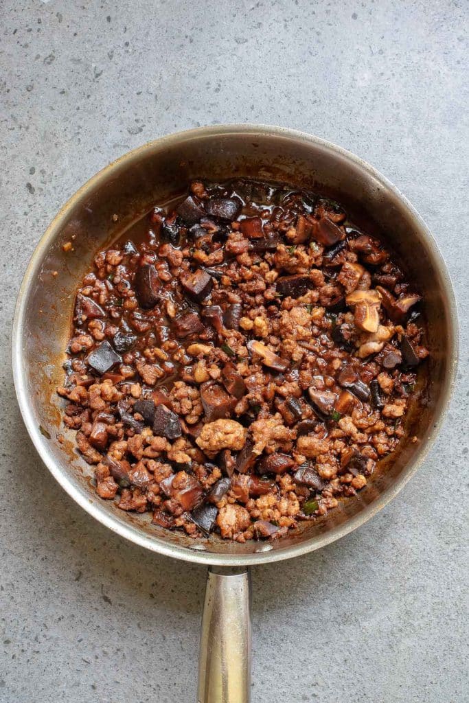 A pan filled with browned minced meat and diced eggplant cooked in a savory sauce. The mixture appears rich and well-seasoned, with a glossy texture, against a grey countertop background.