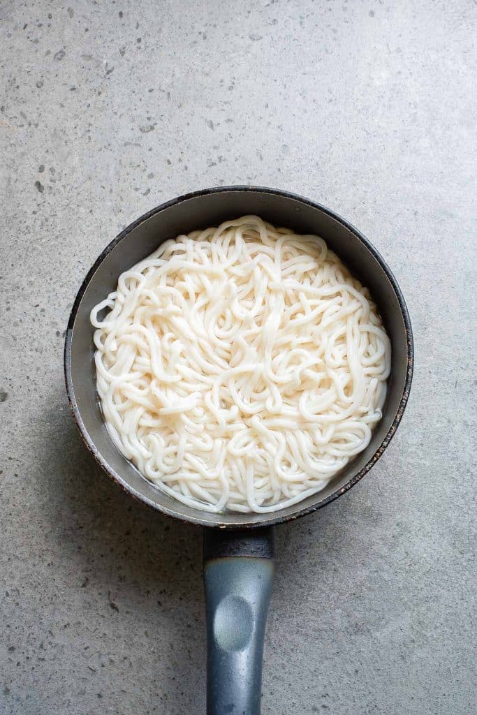 A saucepan filled with cooked white noodles against a gray textured background.