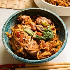 A bowl of stir-fried noodles with chicken, vegetables, and sesame seeds, served in a blue ceramic bowl. Chopsticks are placed beside the bowl on a textured surface.