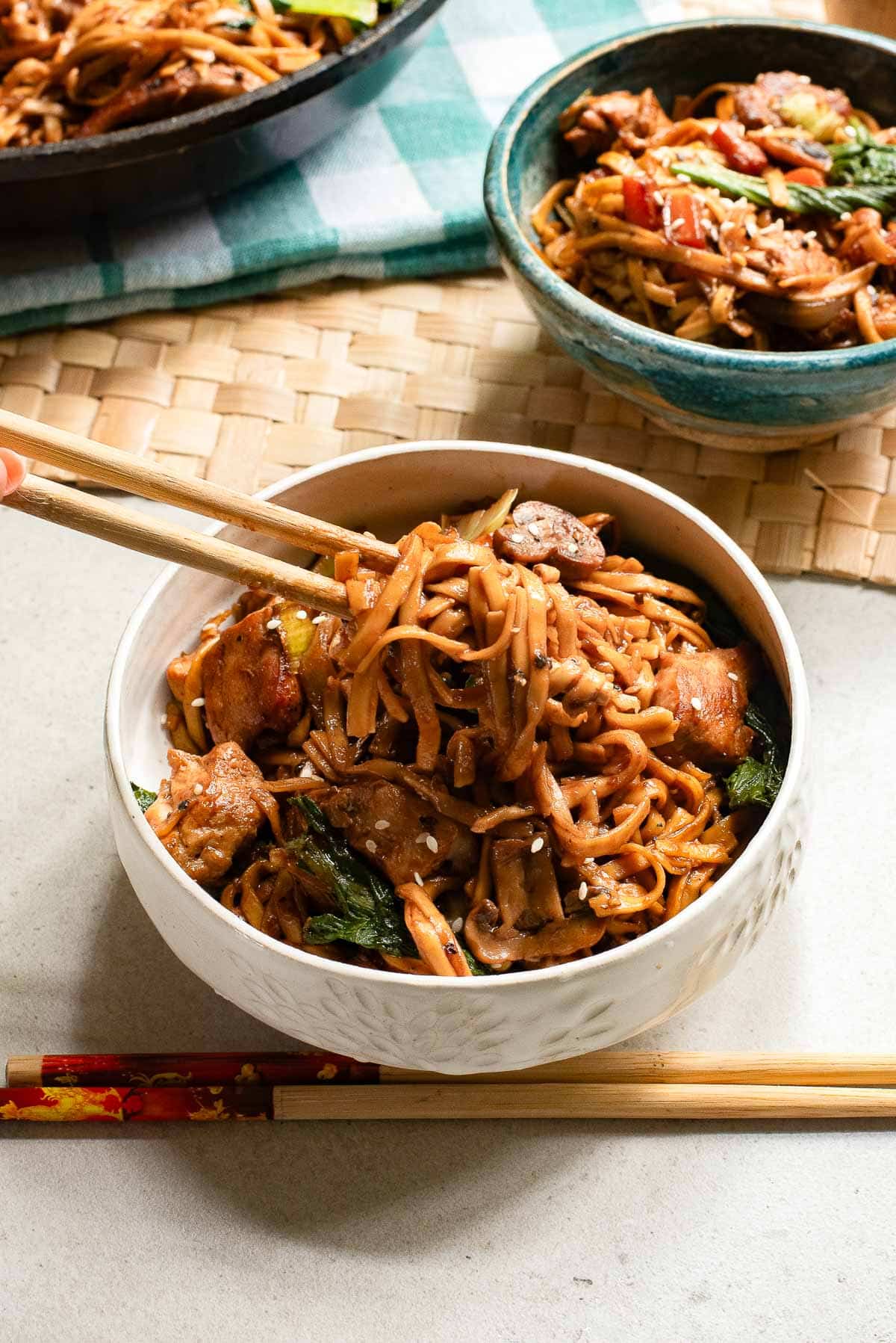 A bowl of stir-fried noodles with vegetables and meat is being held with chopsticks over a white bowl. A second bowl and a frying pan with more noodles are visible in the background. A green and white checkered napkin is underneath.
