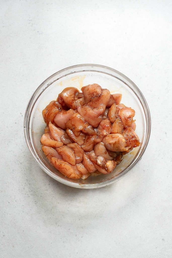 A glass bowl filled with raw marinated chicken pieces on a white countertop. The chicken is cut into bite-sized chunks and appears seasoned with herbs and spices.