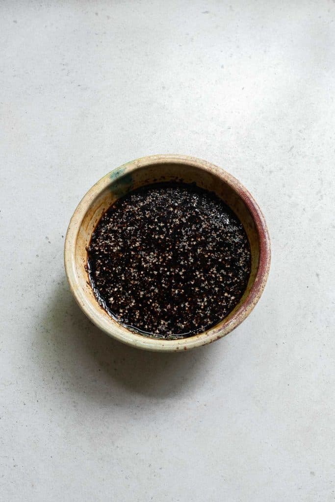 A ceramic bowl filled with a dark, speckled sauce sits on a light, textured surface.