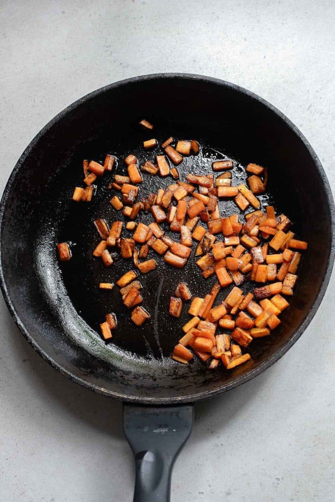 Chunks of carrot sizzling in a black frying pan on a light gray surface. The carrots are browned and seasoned, suggesting they are being sautéed.