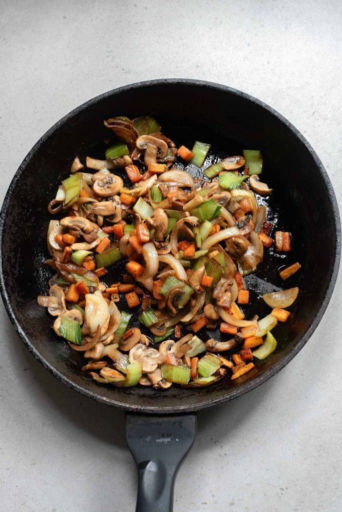 A frying pan with sautéed vegetables including mushrooms, onions, green bell peppers, and carrots on a light gray background.
