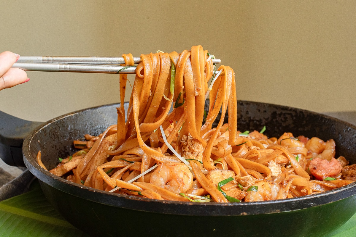 A hand using chopsticks to lift a portion of stir-fried noodles from a pan. The dish includes shrimp, bean sprouts, and chopped green onions, all coated in a savory sauce.