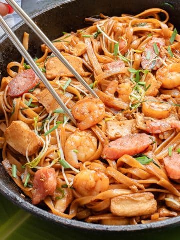 A skillet filled with stir-fried noodles, shrimp, chicken, and sausage, garnished with bean sprouts and chopped green onions. Chopsticks are being used to pick up the noodles.