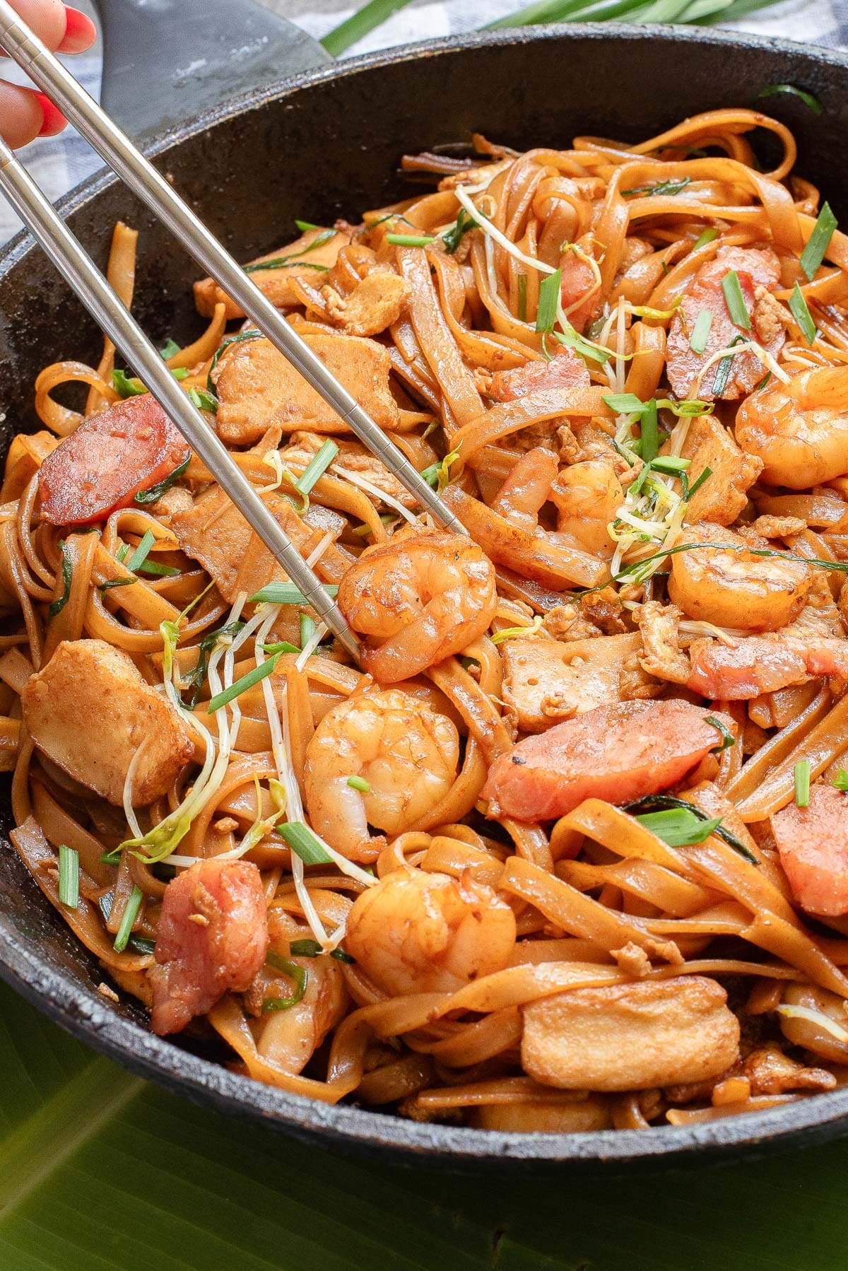 A skillet filled with stir-fried noodles, shrimp, chicken, and sausage, garnished with bean sprouts and chopped green onions. Chopsticks are being used to pick up the noodles.