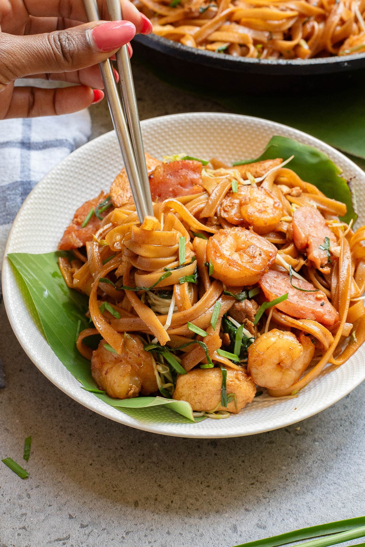 A hand using chopsticks to lift noodles from a bowl of stir-fried noodles with shrimp, sausage, and herbs, served on a banana leaf. A skillet with more noodles is in the background.