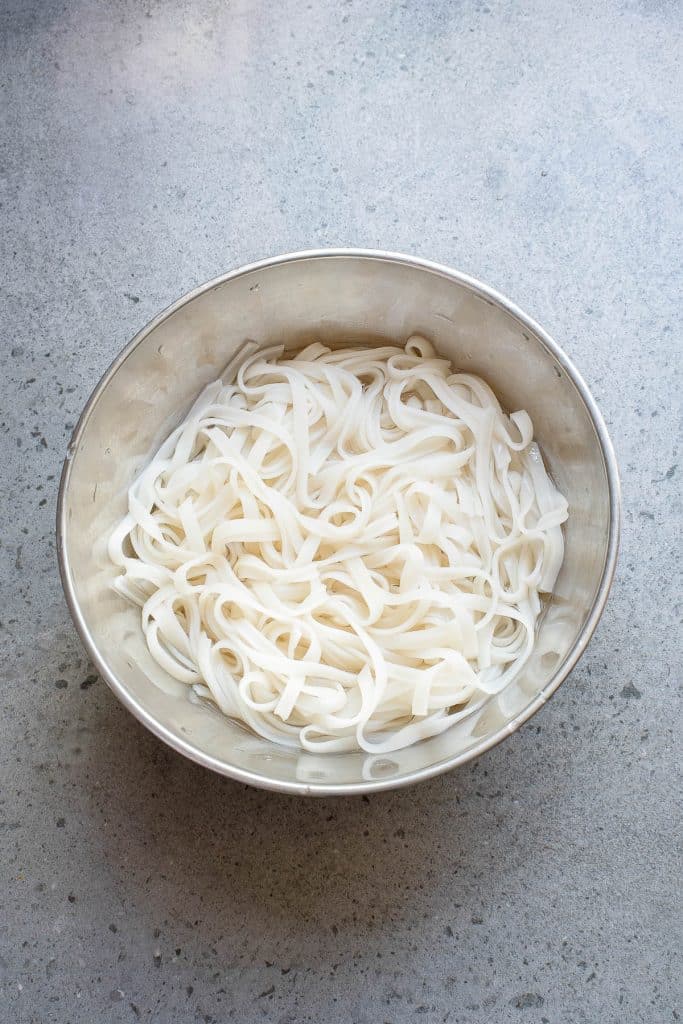 A stainless steel bowl filled with cooked rice noodles on a gray countertop. The noodles are white and arranged in a loose pile, showing their flat and wide shape.