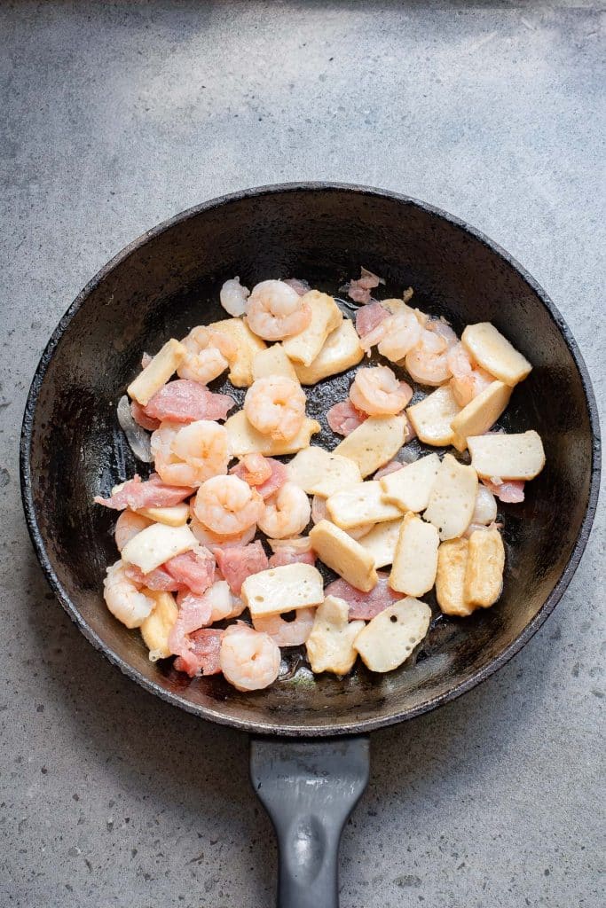 A frying pan containing shrimp and sliced fish cake pieces being cooked on a gray surface.