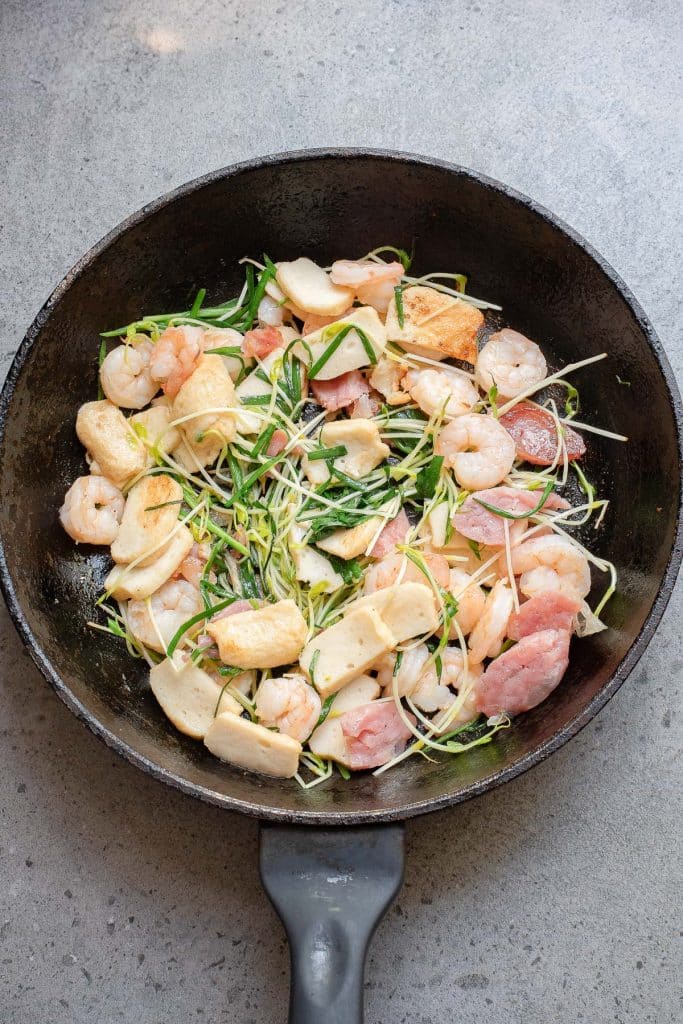A skillet filled with ingredients for a seafood and vegetable stir-fry, including shrimp, sliced mushrooms, chunks of fish, and julienned green onions. The raw ingredients are arranged on a textured gray surface.