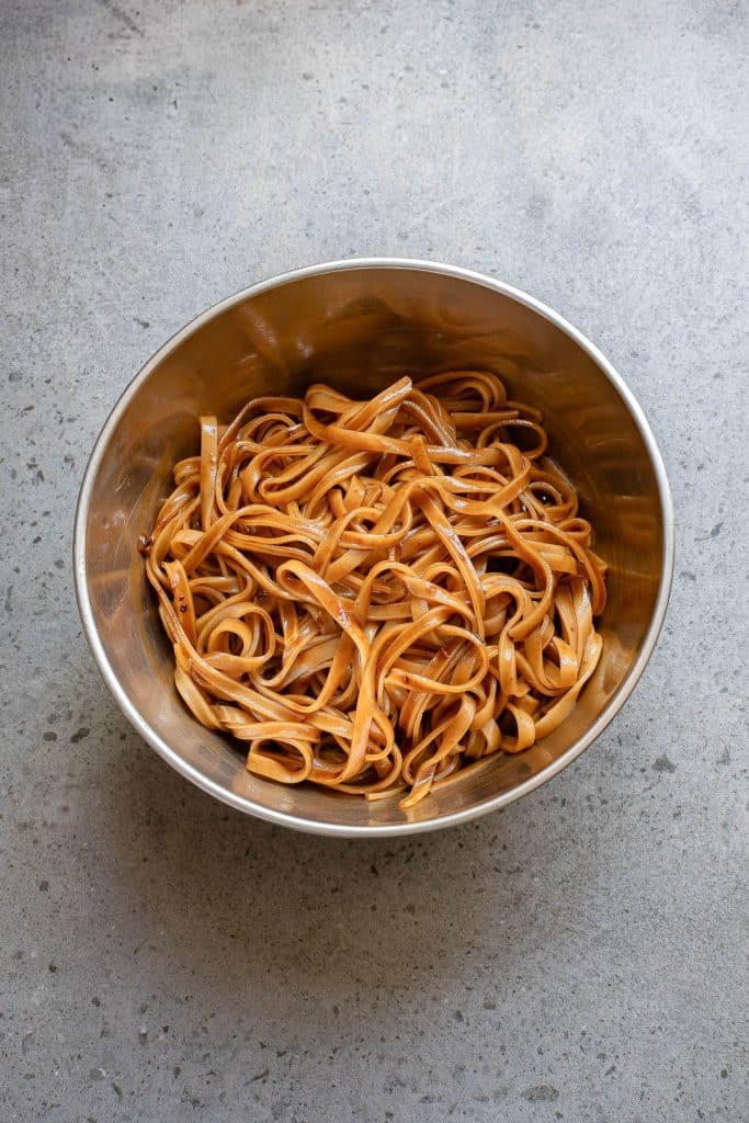 A metal bowl filled with cooked tagliatelle pasta on a gray textured surface.