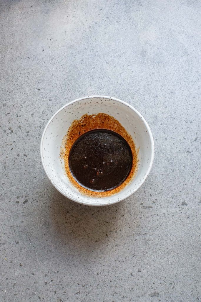 A speckled white bowl contains dark liquid with a frothy edge, resting on a textured gray surface.