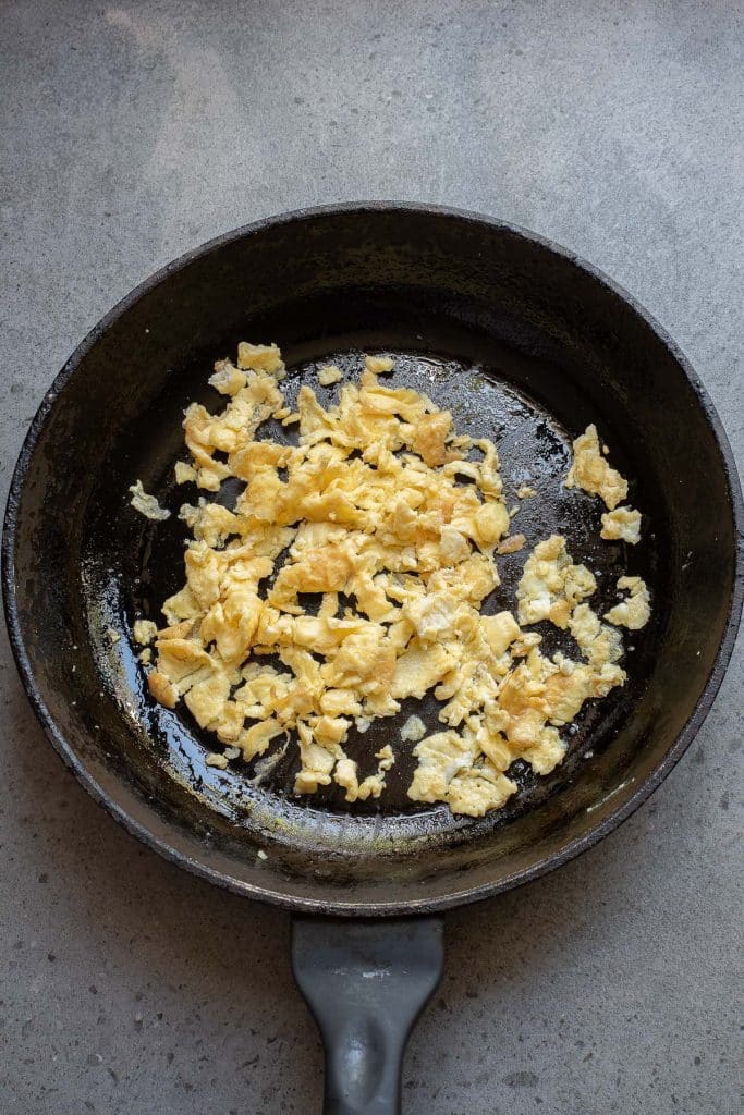 A black skillet containing cooked scrambled eggs sits on a gray surface. The eggs are lightly browned in some areas and have an uneven, crumbly texture. The pan shows slight marks of use.