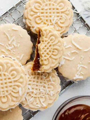 A wire rack holds neatly arranged, decorative cookies dusted with coconut flakes. Some cookies are sandwiched with a caramel filling. A small bowl with caramel is nearby on a white marble surface.