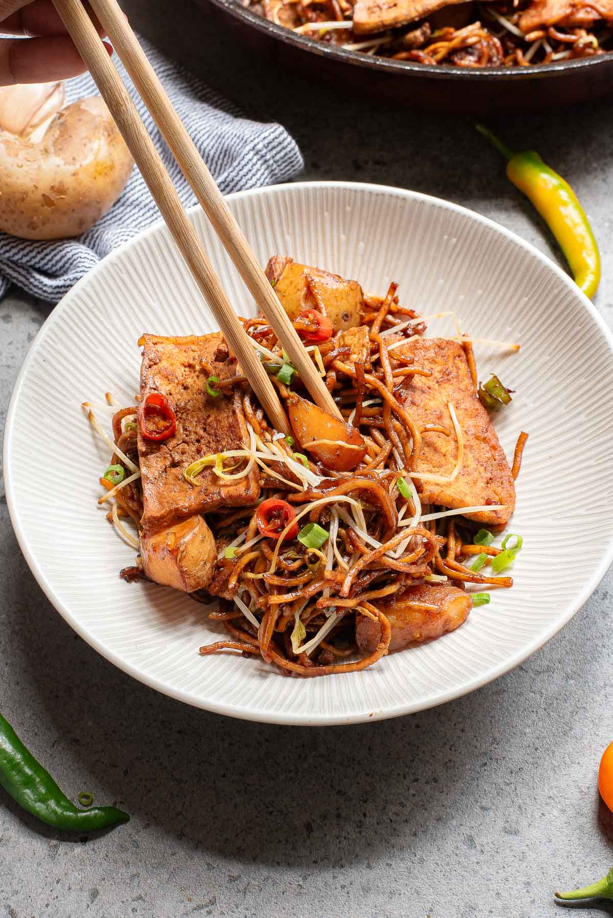 A bowl of stir-fried noodles is topped with crispy tofu, bean sprouts, sliced red chilies, and chopped green onions. A pair of chopsticks hovers over the dish. Fresh peppers and an onion are visible on the table.