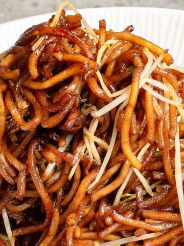 A close-up of a plate of noodles with a glossy brown sauce, garnished with bean sprouts. A fork is lifting a portion of the noodles. The background is a textured surface and a striped cloth is partially visible.