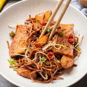 A plate of stir-fried noodles with tofu, bean sprouts, and scallions, garnished with sliced red chili. Two chopsticks rest on the side of the dish.