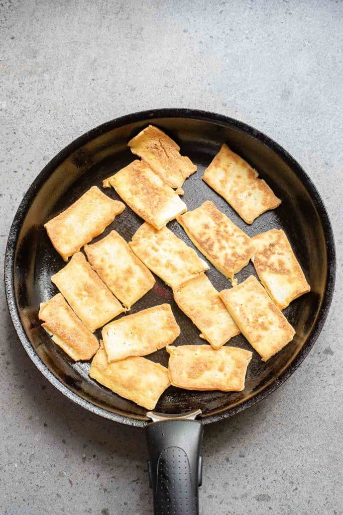 A frying pan on a gray surface contains golden-brown, rectangular pieces of fried tofu.