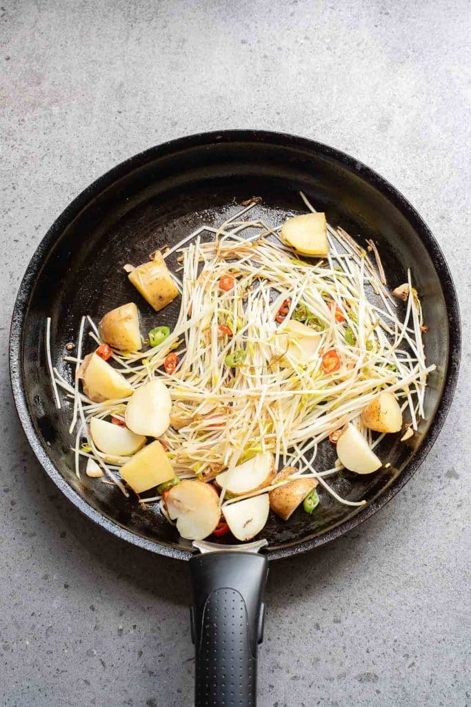 A frying pan containing chopped potatoes, bean sprouts, sliced red chili peppers, and green onions is placed on a gray countertop. The ingredients appear to be in the initial stages of cooking.
