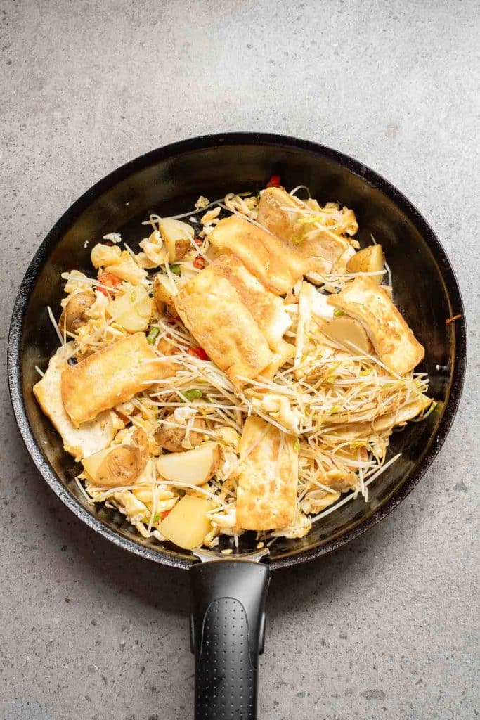 A frying pan filled with sautéed bean sprouts, tofu slices, and chunks of vegetables, including cabbage and potatoes, on a gray countertop.