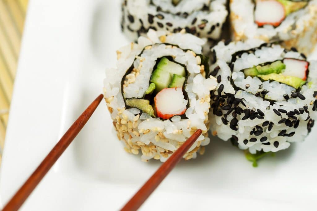 Close-up of sushi rolls on a white plate. The rolls are filled with crab stick, cucumber, and avocado, and topped with black and white sesame seeds. A pair of wooden chopsticks is positioned near the rolls.