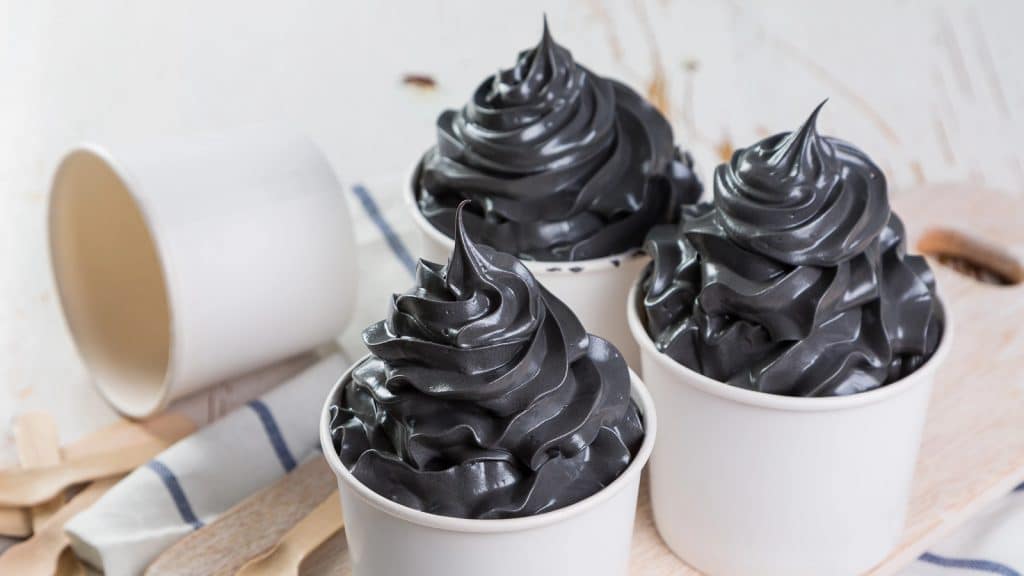 Three cups of dark charcoal-colored soft serve ice cream are arranged on a light wooden surface. An empty cup and wooden sticks lie beside them, with a striped cloth partially visible in the background.