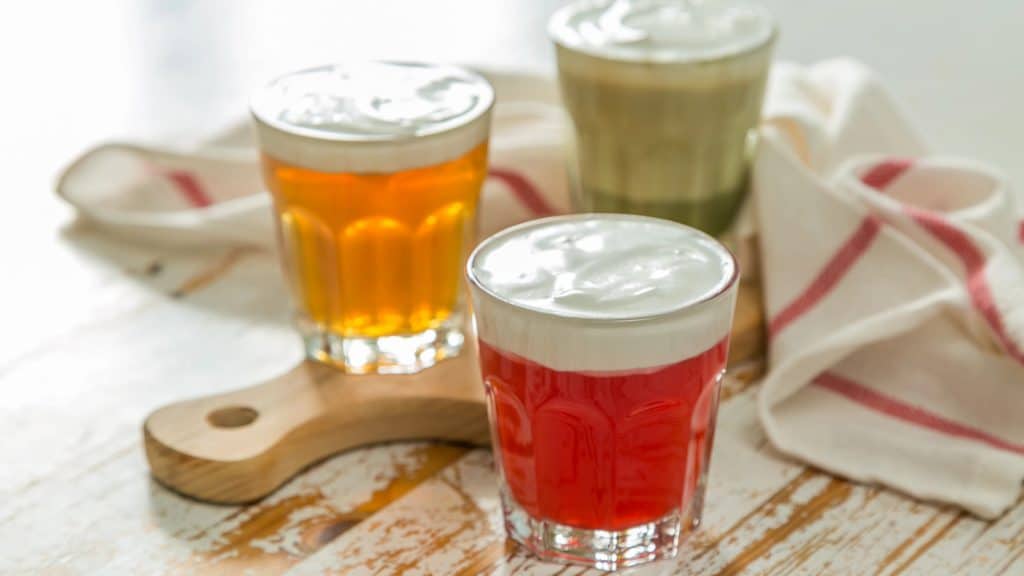 Three glasses of colorful drinks topped with frothy cream sit on a wooden serving board. The drinks are orange, green, and red. A white cloth with red stripes is partially visible in the background on a wooden table.