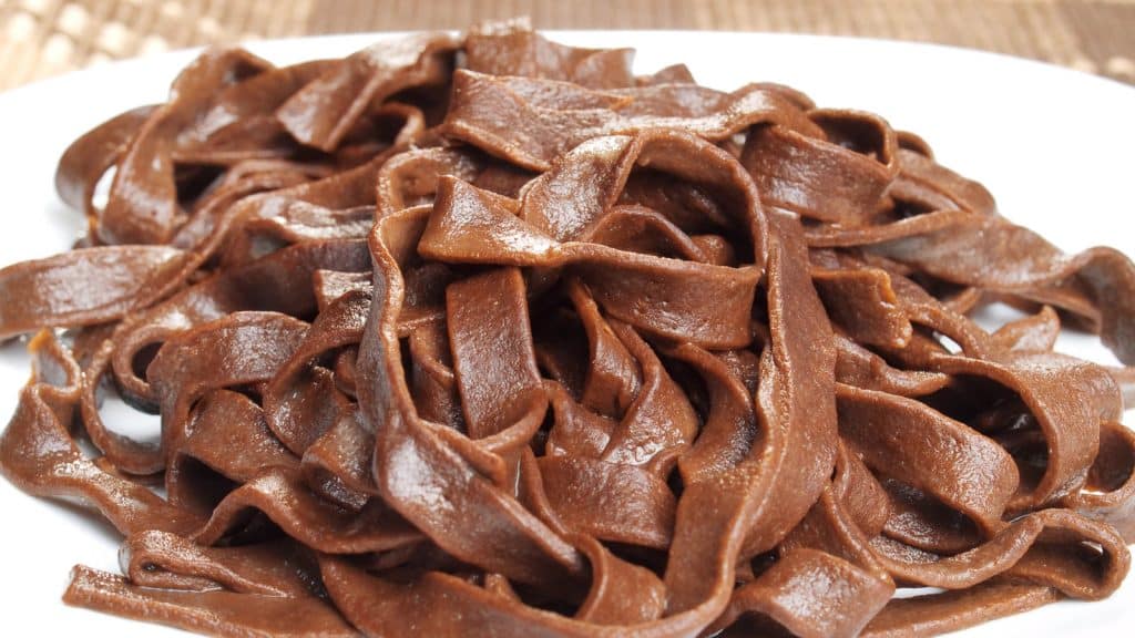 A close-up of chocolate pasta ribbons arranged on a white plate. The pasta is glossy and rich in color, with a few folds and twists visible. The background is softly blurred, enhancing the focus on the pasta.
