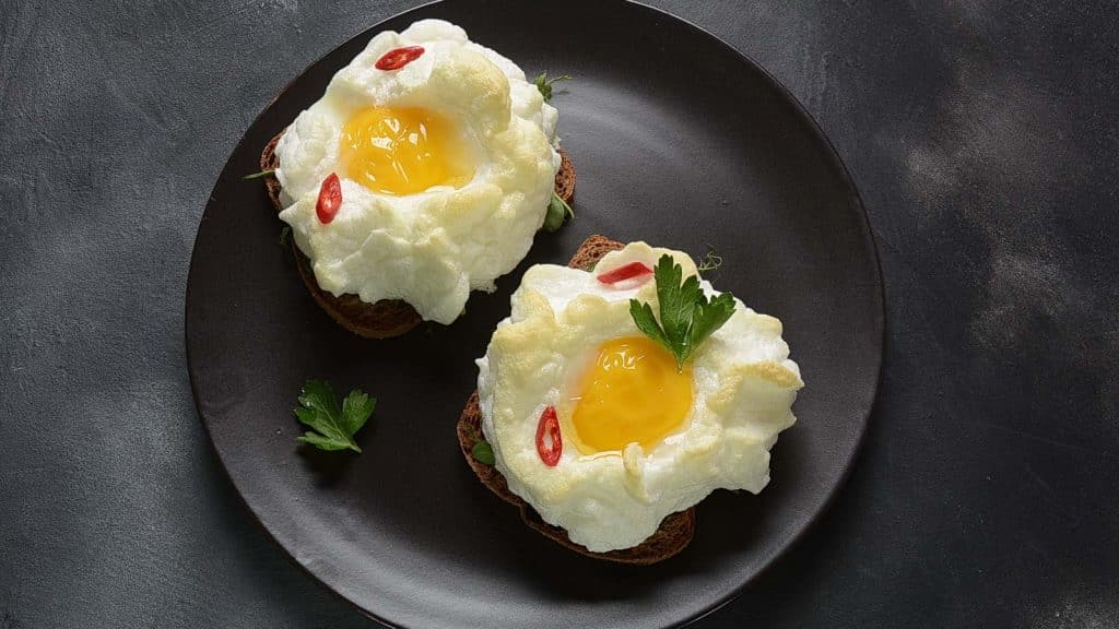 Two cloud egg toasts sit on a black plate. Each features a fluffy baked egg white with a yolk on top of toast, garnished with parsley and red pepper slices. The background is a textured gray surface.