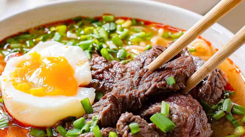 Close-up of a bowl of ramen with chopsticks holding slices of beef. The dish is topped with a soft-boiled egg, green onions, and immersed in a rich, reddish broth.