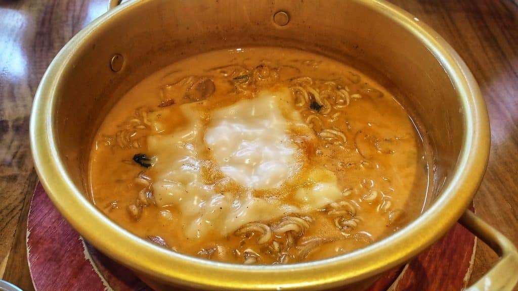 A pot of spicy ramen with noodles, broth, and melted cheese on top. The pot is set on a wooden table.