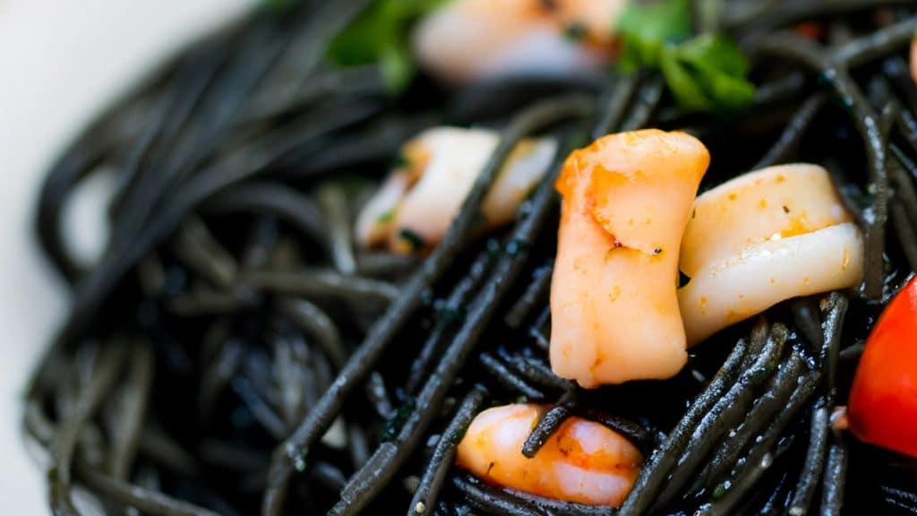 Close-up of black spaghetti garnished with pieces of seafood and herbs. A cherry tomato is visible on the side, adding a pop of color to the dish.