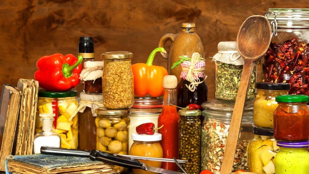 A variety of jars filled with preserved foods, such as olives, peppers, and spices, are arranged on a wooden surface. A wooden spoon rests against one jar, surrounded by old books and kitchen utensils. Colorful bell peppers add a fresh touch.