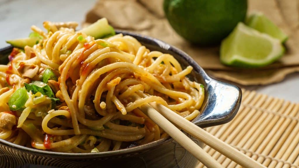 A bowl of peanut noodles garnished with chopped peanuts and green onions, with a pair of chopsticks resting on the side. Lime wedges and a whole lime are in the background on a bamboo mat.