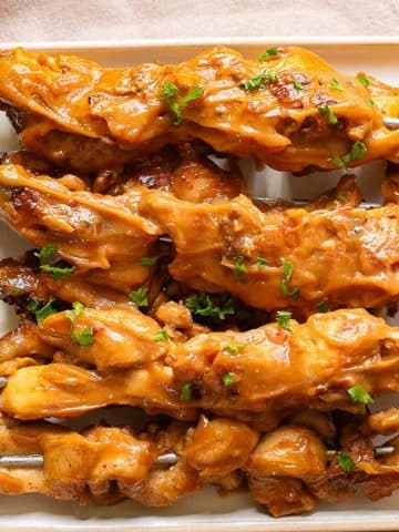 Five skewers of grilled chicken satay are arranged on a rectangular white plate, garnished with chopped herbs. The chicken is coated in a rich, golden-brown peanut sauce, providing a savory and appetizing appearance.