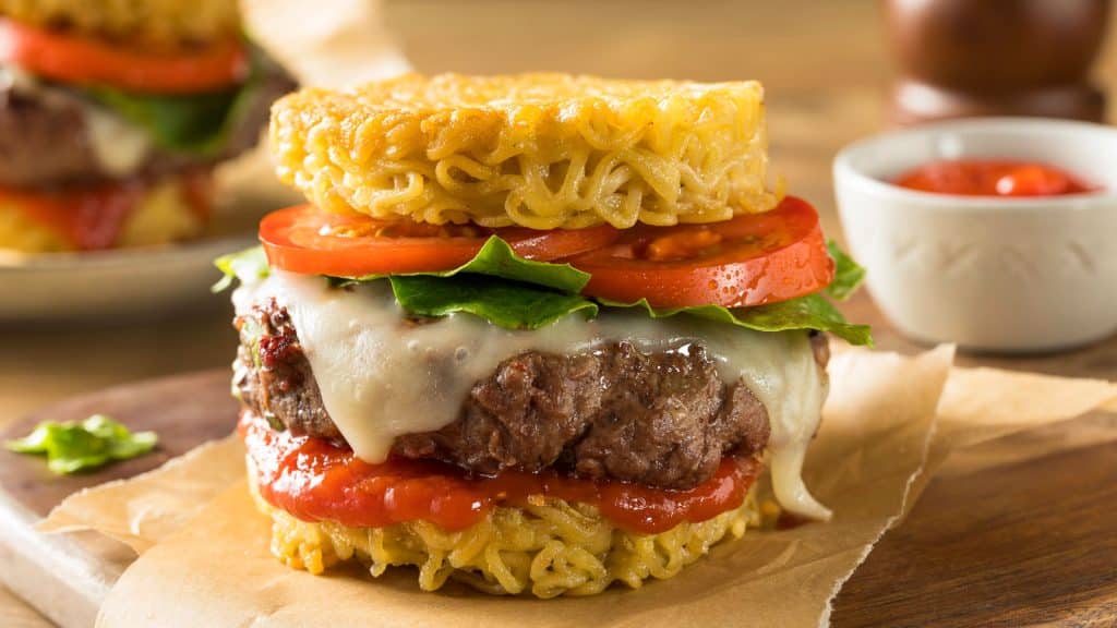 A creative ramen burger with crispy ramen noodle buns, a juicy beef patty, melted cheese, fresh lettuce, tomato slices, and ketchup. It is presented on parchment paper with a small bowl of ketchup in the background.