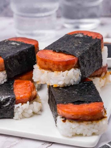 Four pieces of Spam musubi, consisting of grilled Spam on rice wrapped with seaweed, are neatly arranged on a white plate. A can of Spam, a bowl of sauce, and a wooden bowl are in the background.