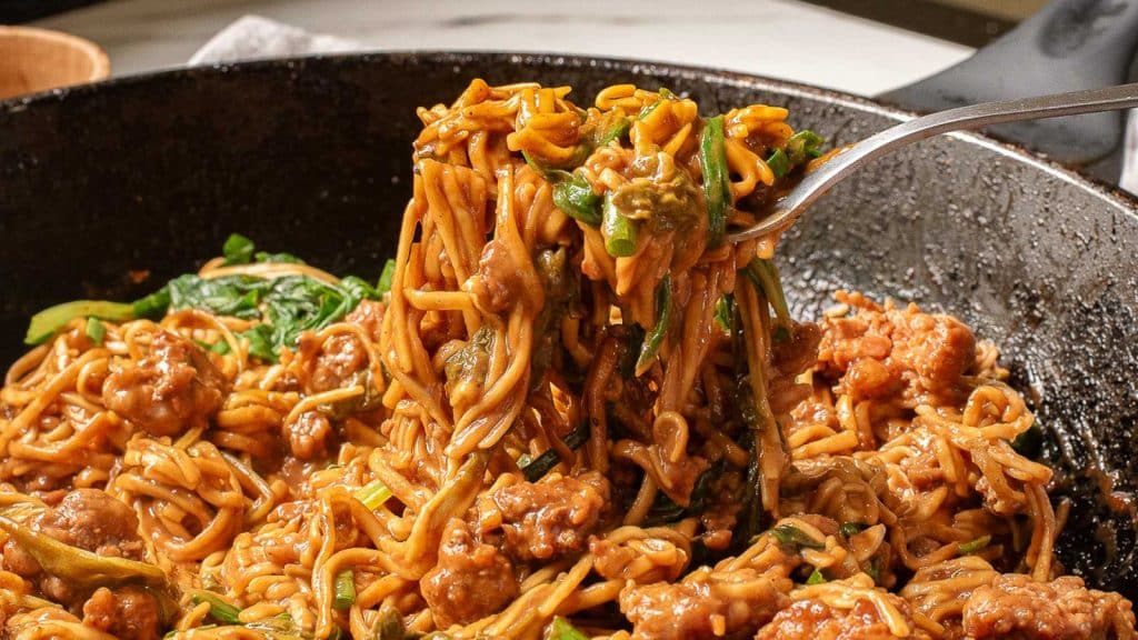 Close-up of a skillet containing a savory noodle dish with ground meat and green vegetables. A fork is lifting a portion of the noodles, showcasing the sauce and ingredients in the mix.