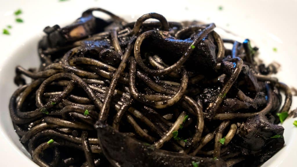 A close-up of black squid ink pasta with pieces of squid and a garnish of chopped parsley, served on a white plate.