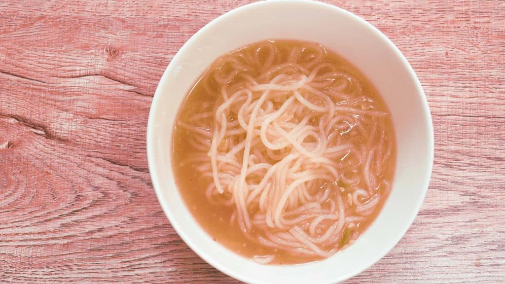A white bowl filled with noodles in clear broth, placed on a wooden surface.