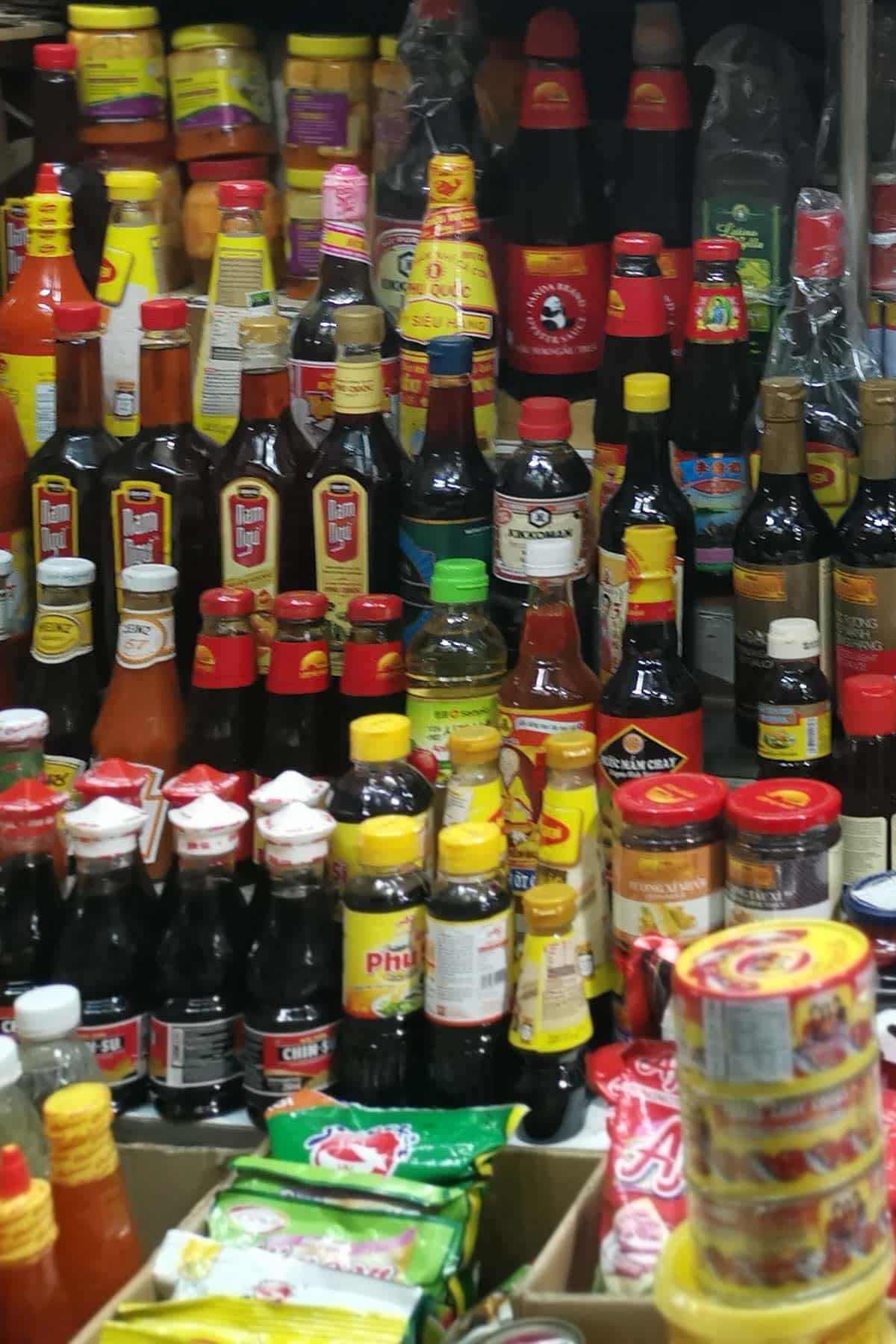 A crowded store shelf filled with various bottles and containers of sauces, seasonings, and condiments, including soy sauce, chili sauce, and ketchup, along with packets and jars, all in different shapes, colors, and sizes.