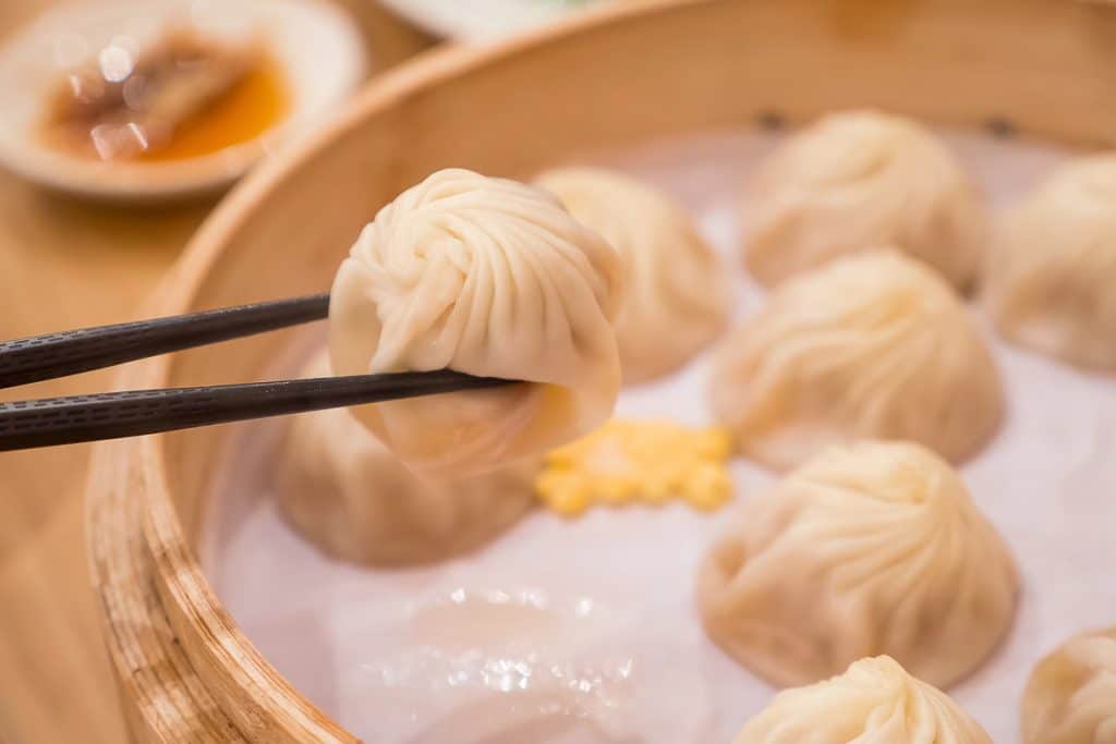Chopsticks holding a steamed dumpling above a bamboo steamer filled with similar dumplings. A small dish with sauce is visible in the background.