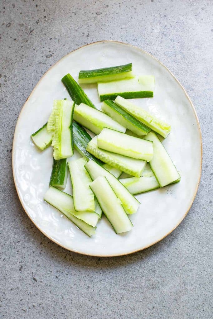 Sliced cucumber sticks arranged on a round, white ceramic plate set against a gray textured background.