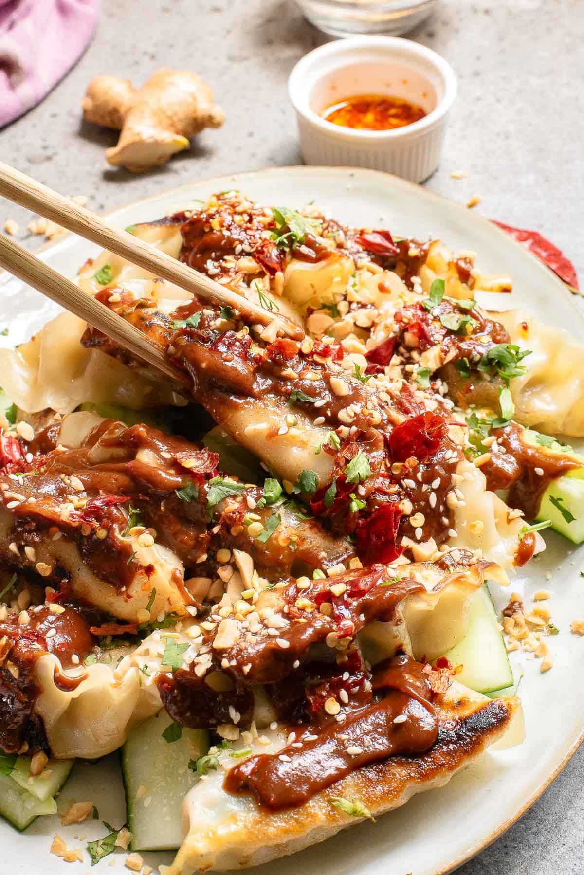 A plate of dumplings drizzled with chili sauce, garnished with chopped cilantro and crushed peanuts. Chopsticks are holding one dumpling. Slices of cucumber are visible under the dumplings. A small bowl of dipping sauce is in the background.