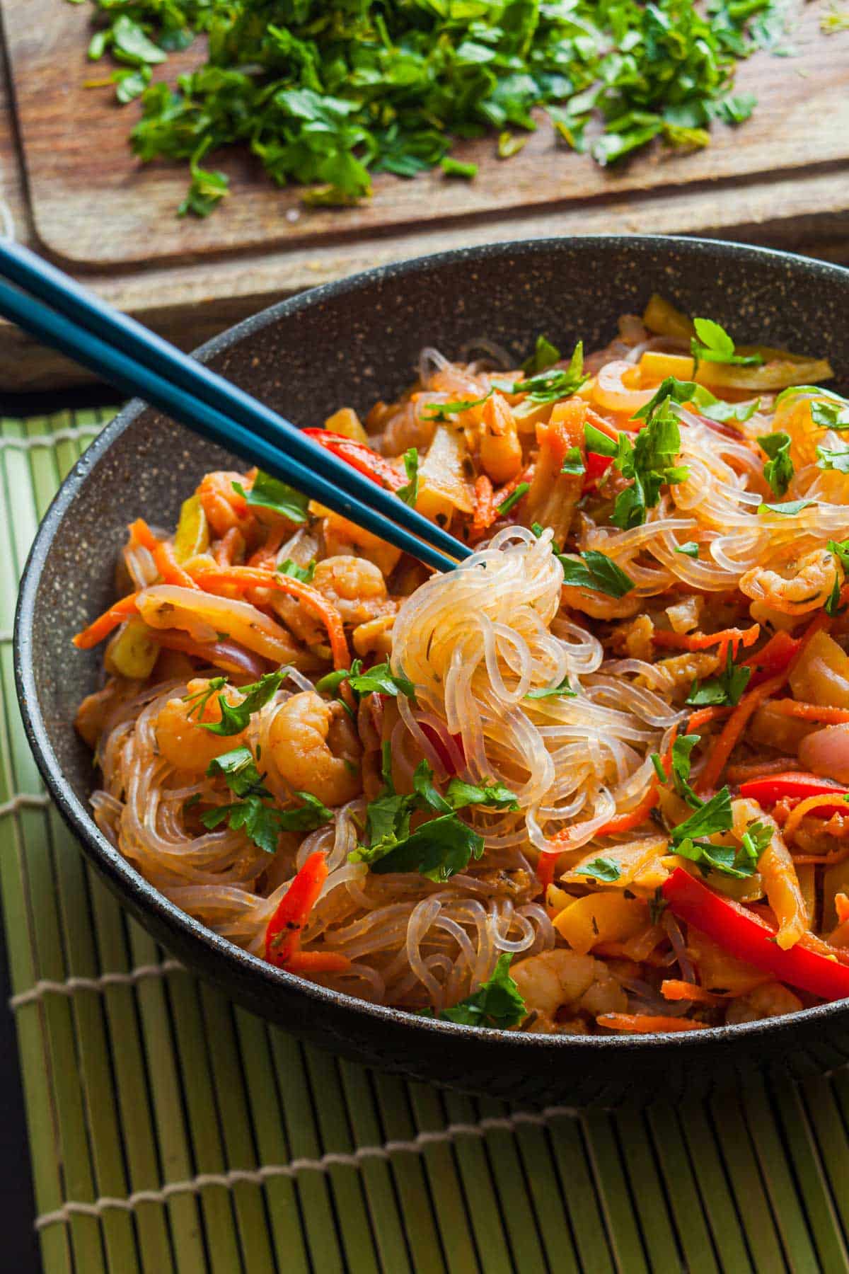 A bowl of stir-fried glass noodles with vegetables and herbs is served with chopsticks. The dish is garnished with chopped cilantro, and in the background, more cilantro is visible on a chopping board.