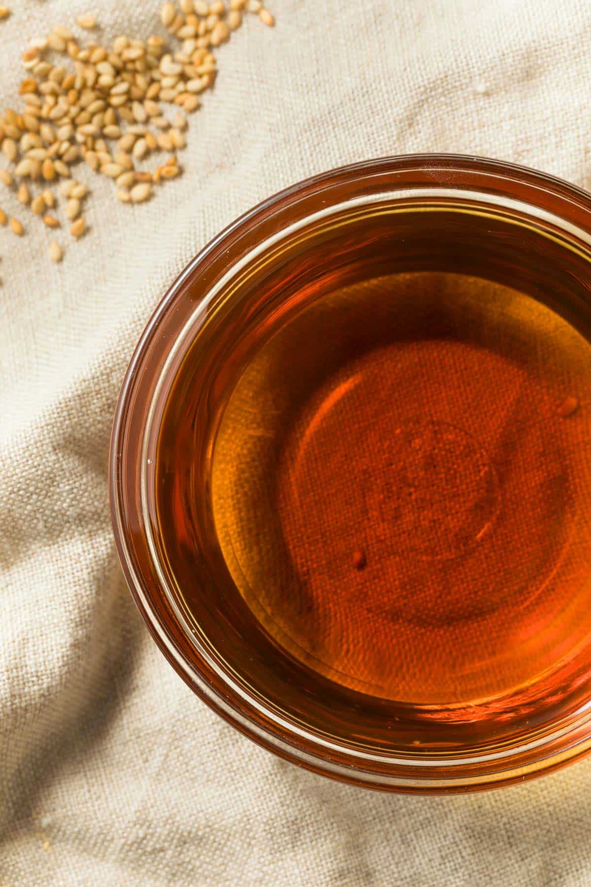 A glass bowl of sesame oil sits on a textured, beige fabric surface. A small pile of sesame seeds is visible in the upper left corner. The oil has a rich, amber color.
