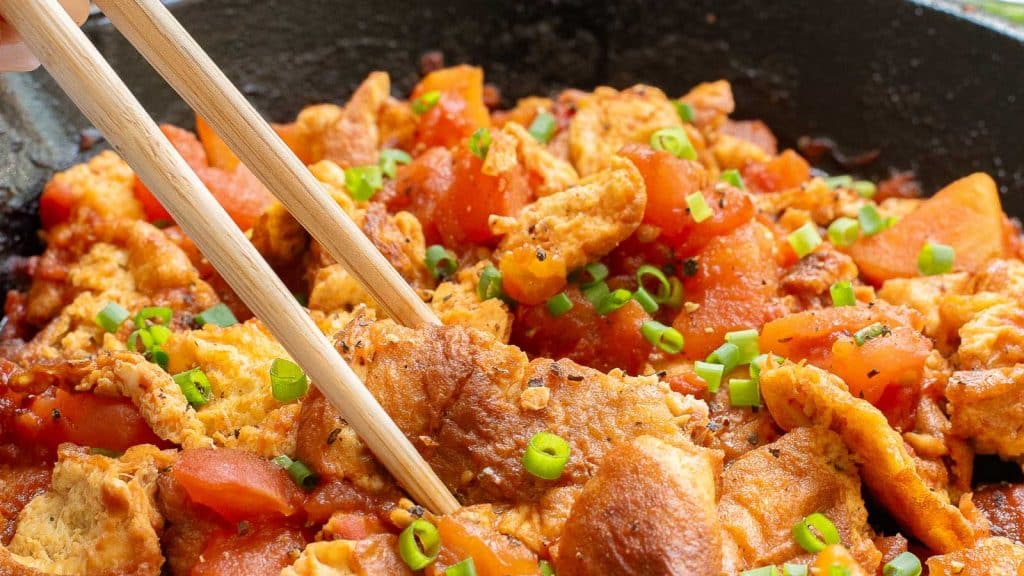 Close-up of a dish featuring succulent pieces of cooked tofu with diced tomatoes, garnished with chopped green onions. A pair of chopsticks is picking up a tofu piece from a sizzling pan.
