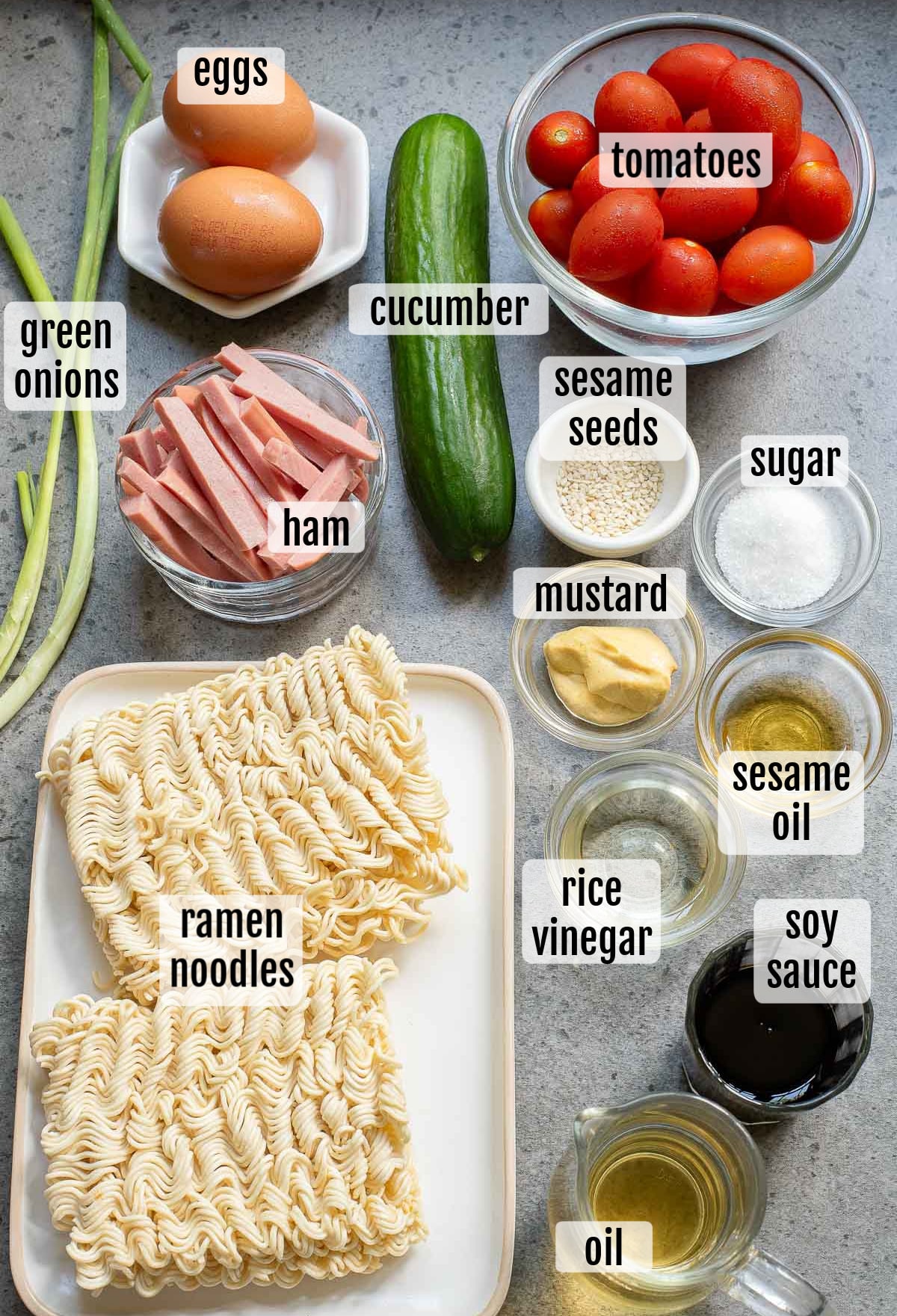 Ingredients for a hiyashi chuka dish are artfully arranged on a surface, featuring labeled eggs, tomatoes, cucumber, green onions, ham, sesame seeds, mustard, sugar, sesame oil, rice vinegar, soy sauce, oil and ramen noodles.